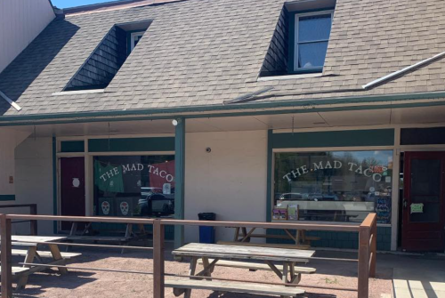 Exterior of "The Mad Taco" restaurant with picnic tables and large windows, featuring a sloped roof and colorful signage.