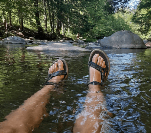 Feet in sandals submerged in a calm river, surrounded by rocks and lush greenery.