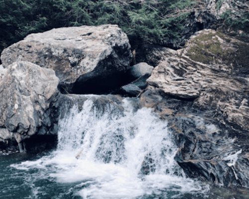 A small waterfall cascades over rocky terrain, surrounded by greenery and natural stone formations.