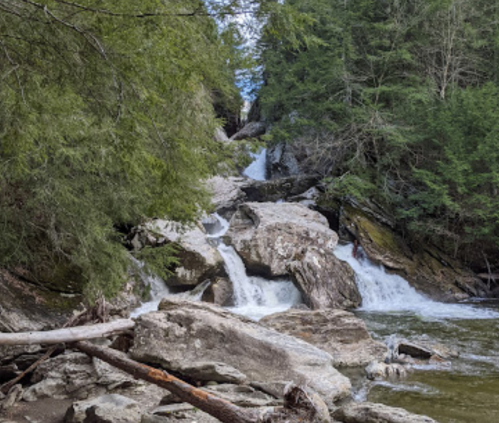 A serene waterfall cascades over rocks, surrounded by lush green trees and a calm river.