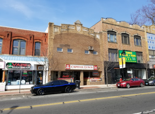 A street view featuring brick buildings, including Capitol Lunch, with parked cars and clear blue skies.