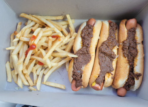 Three hot dogs topped with chili in a box, accompanied by a side of French fries and ketchup.
