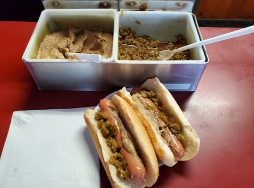 Three hot dogs in buns topped with relish, next to containers of mustard and chopped onions on a red table.