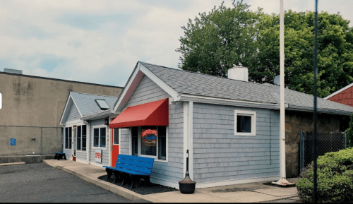 A small, charming building with a blue bench and red awning, surrounded by greenery and a cloudy sky.