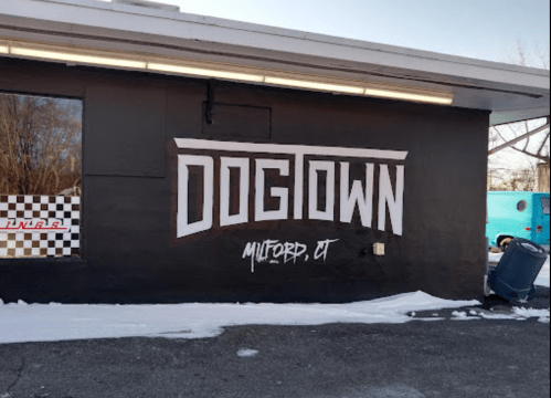 Mural on a black wall reading "DOGTOWN, MILFORD, CT" with snow on the ground and a trash can nearby.