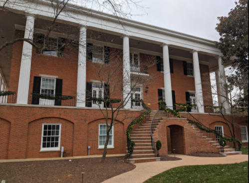 A brick building with white columns, large windows, and a staircase surrounded by greenery.