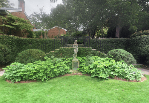 A garden scene featuring a statue surrounded by lush greenery and low shrubs, with a brick border and a hedge backdrop.