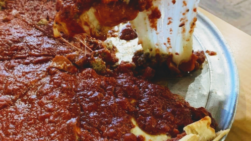 A slice of deep-dish pizza being lifted, showcasing gooey cheese and a rich tomato sauce on a metal pizza pan.