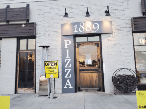 Exterior of 1889 Pizza with a welcoming entrance, open sign, and stacked firewood beside the door.