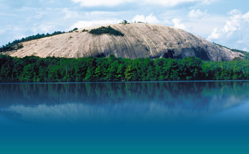 A large, rocky hill reflects in a calm lake, surrounded by lush green trees under a blue sky with clouds.