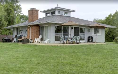 A modern house with a brick chimney, patio furniture, and umbrellas, surrounded by a green lawn and trees.