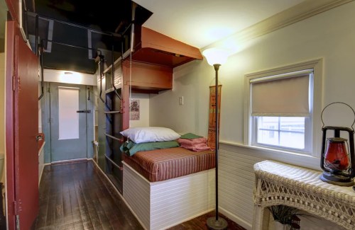 Cozy room with a bed, shelves, a lamp, and a door leading to another space, featuring wooden floors and light walls.