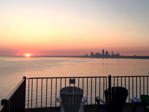 Sunset over a calm lake with a city skyline in the distance, featuring two empty chairs in the foreground.