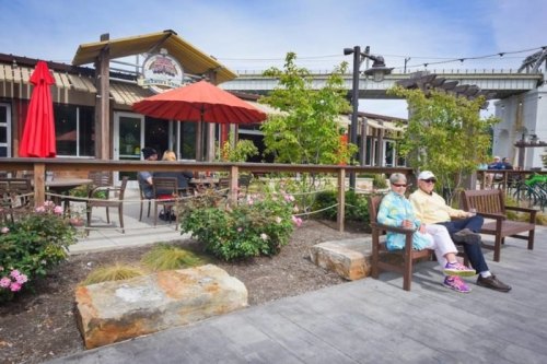 Two people sit on a bench outside a restaurant, surrounded by greenery and flowers, with umbrellas providing shade.