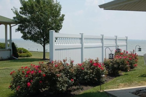 A white fence borders a garden with blooming roses, overlooking a calm lake under a clear sky.