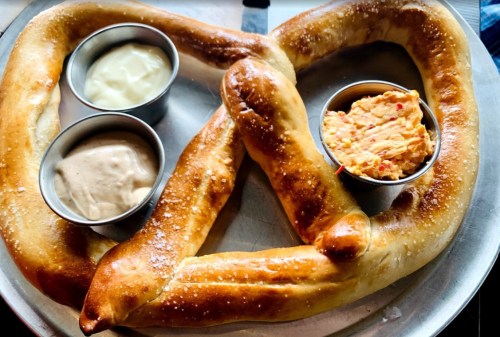 A large pretzel on a metal tray, served with three dipping sauces in small cups.