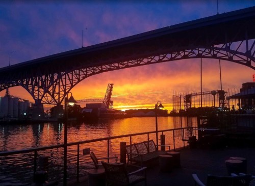 A vibrant sunset casts colorful hues over a river, framed by a bridge and silhouettes of nearby structures.