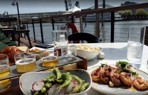 A riverside dining scene featuring shrimp, edamame, popcorn, and craft beers on a sunny day.