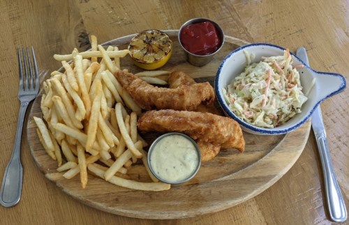 A wooden platter with crispy fish fillets, fries, coleslaw, a lemon wedge, and dipping sauces.
