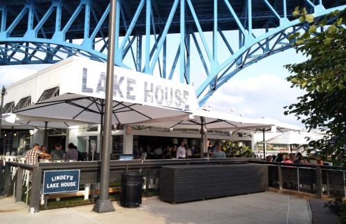Exterior of Lindey's Lake House, featuring outdoor seating under umbrellas and a blue bridge overhead.
