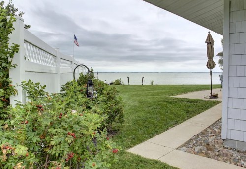 A serene lakeside view with a grassy area, white fence, and cloudy sky, featuring a flag and a patio umbrella.