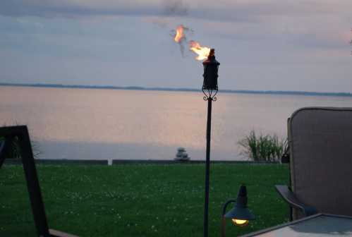 A tiki torch flames beside a calm lake at dusk, with grass and patio furniture in the foreground.