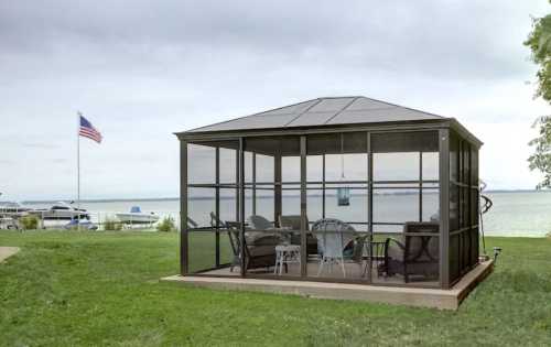 A screened gazebo on a grassy shore, overlooking a lake with boats and an American flag in the background.