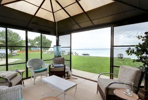 A cozy screened porch with seating, overlooking a calm lake and green lawn.