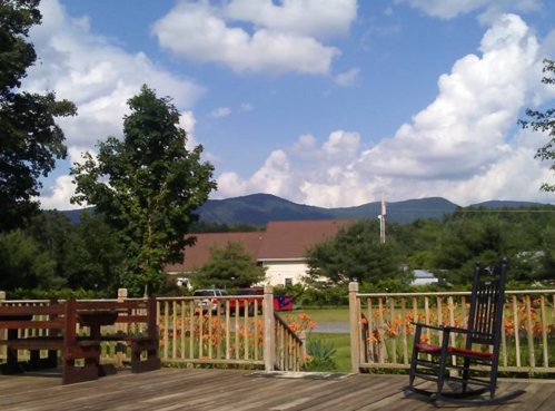 A scenic view of mountains under a blue sky with fluffy clouds, framed by trees and a wooden deck with a rocking chair.