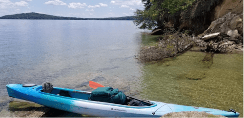 A blue kayak rests on a rocky shore by calm water, surrounded by trees and a clear sky.
