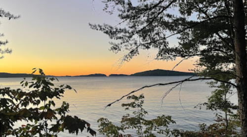 A serene lakeside view at sunset, with silhouettes of trees and distant islands against a colorful sky.