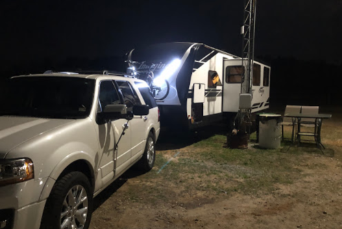 A white SUV parked next to a travel trailer at night, illuminated by bright lights.