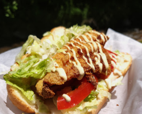 A sandwich with fried fish, lettuce, tomato, and drizzled sauce on a toasted bun, served on parchment paper.