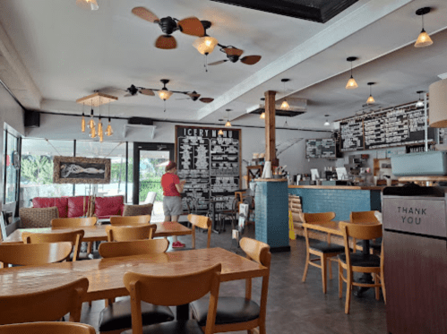 Cozy café interior with wooden tables, chairs, a menu board, and a customer browsing. Bright and inviting atmosphere.