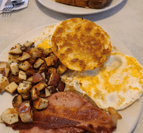 A plate of breakfast featuring two eggs, crispy bacon, seasoned potatoes, and a toasted English muffin.
