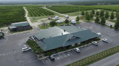 Aerial view of a large building surrounded by parking and fields, with trees and vehicles nearby.