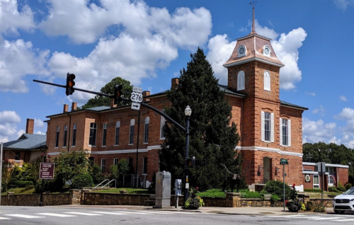 Historic brick building with a clock tower, surrounded by greenery and under a blue sky with fluffy clouds.