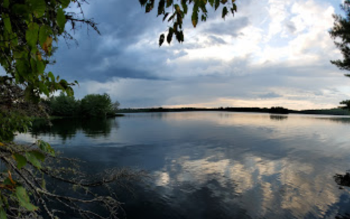 A serene lake scene with calm waters reflecting clouds and trees under a partly cloudy sky.