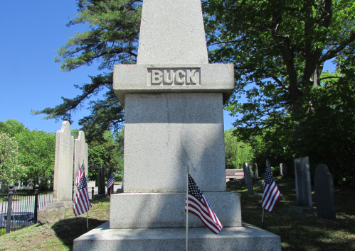 A granite monument with the name "BUCK" engraved, surrounded by small American flags in a cemetery.