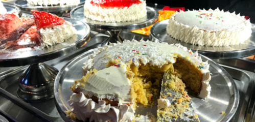 A variety of decorated cakes on silver platters, with one cake partially sliced to reveal its yellow interior.