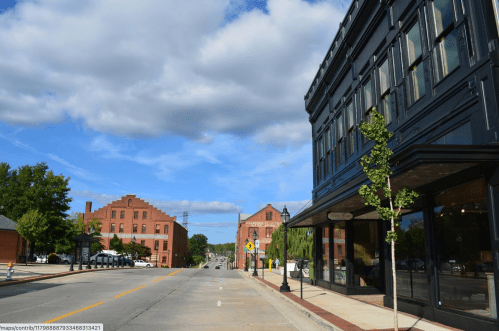 A quiet street scene featuring a historic building, trees, and a blue sky with scattered clouds.