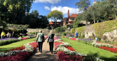 Two people walk through a vibrant garden with colorful flower beds and a historic building in the background.