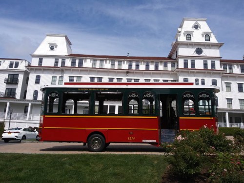 A red and green trolley bus parked in front of a large white historic hotel under a blue sky.