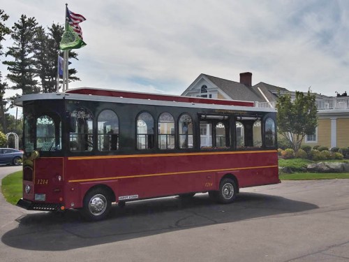 A red and black trolley bus parked near a large yellow house with trees and flags in the background.