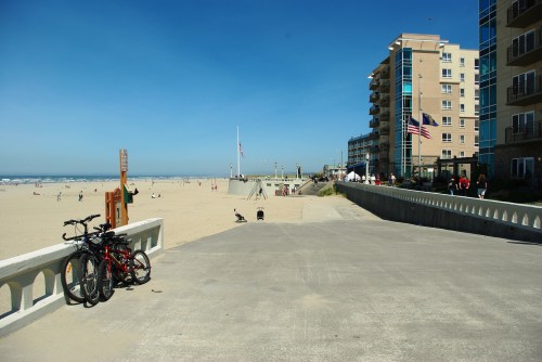 A sunny beach scene with a bike rack, sandy shore, and buildings in the background under a clear blue sky.