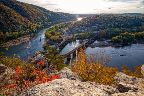 A scenic view of a river winding through autumn-colored hills, with a bridge and a small town in the distance.