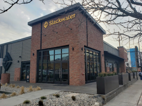 Exterior of a brick building with large windows, featuring the sign "Slackwater" and a landscaped area in front.