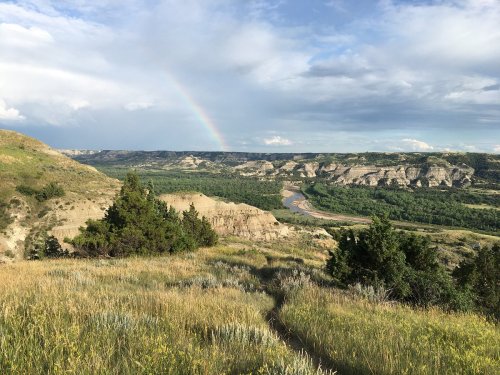 A scenic view of rolling hills and a river, with a rainbow arching across a partly cloudy sky.