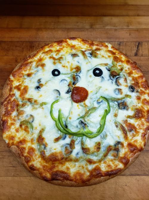 A pizza with a face made of cheese, olives, green peppers, and a pepperoni nose, set on a wooden surface.