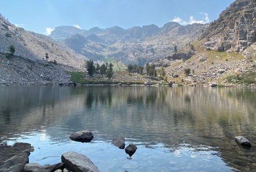 A serene mountain lake reflecting rocky cliffs and greenery under a clear blue sky.